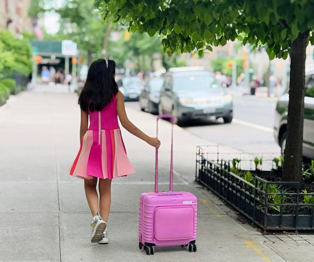 A young girl walks down a city street with a pink suitcase.