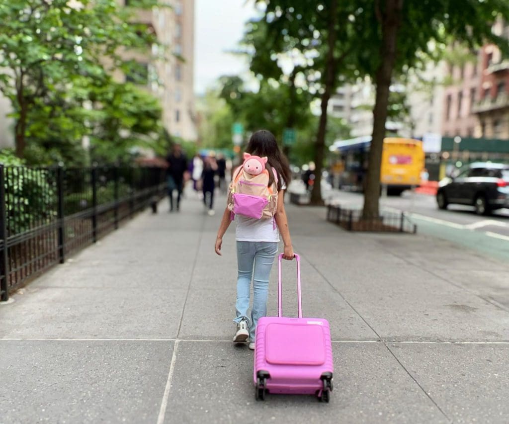 A young girl pulls a suitcase through a city street, while also wearing a backpack.