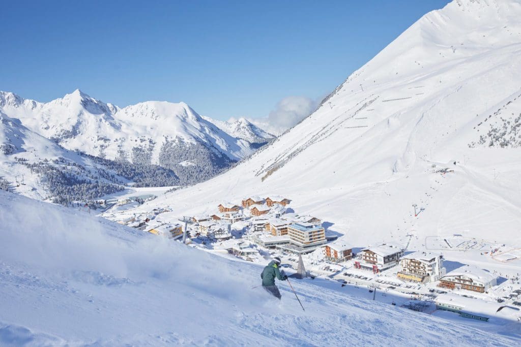 An aerial view of Kühtai, nestled in the snowy mountains of Austria, someone skis past on a snowy trail, knowing about the city is a key part of this family guide to skiing in Kühtai this winter.