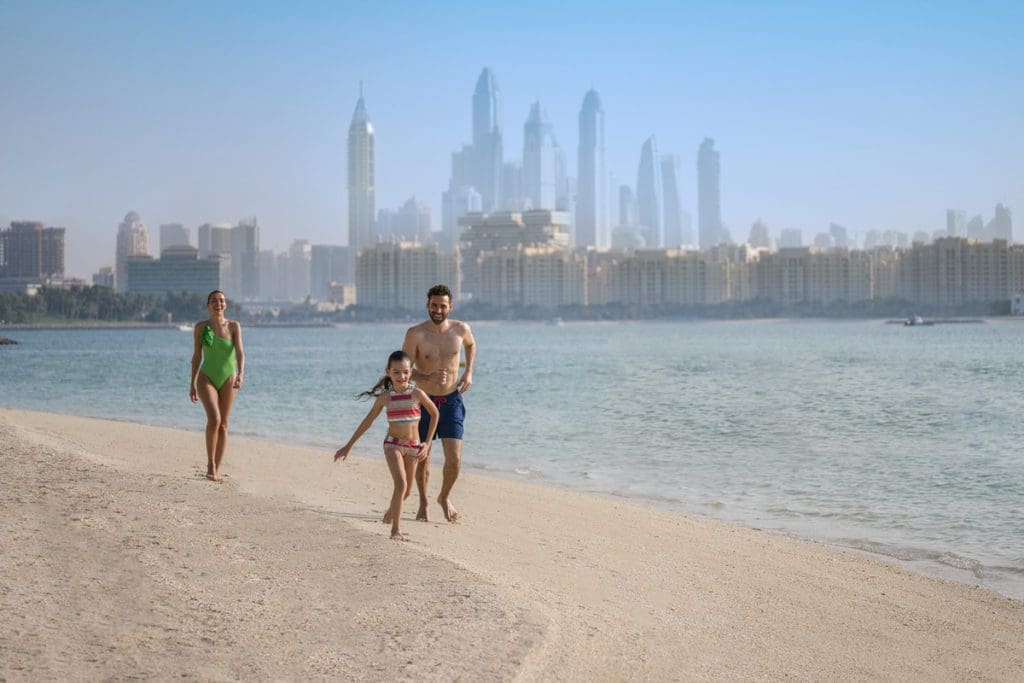 A family of three runs across the sand, with a Dubai skyline in the distance, at Rixos The Palm Hotel and Suites, one of the best beachfront hotels in Dubai for families.
