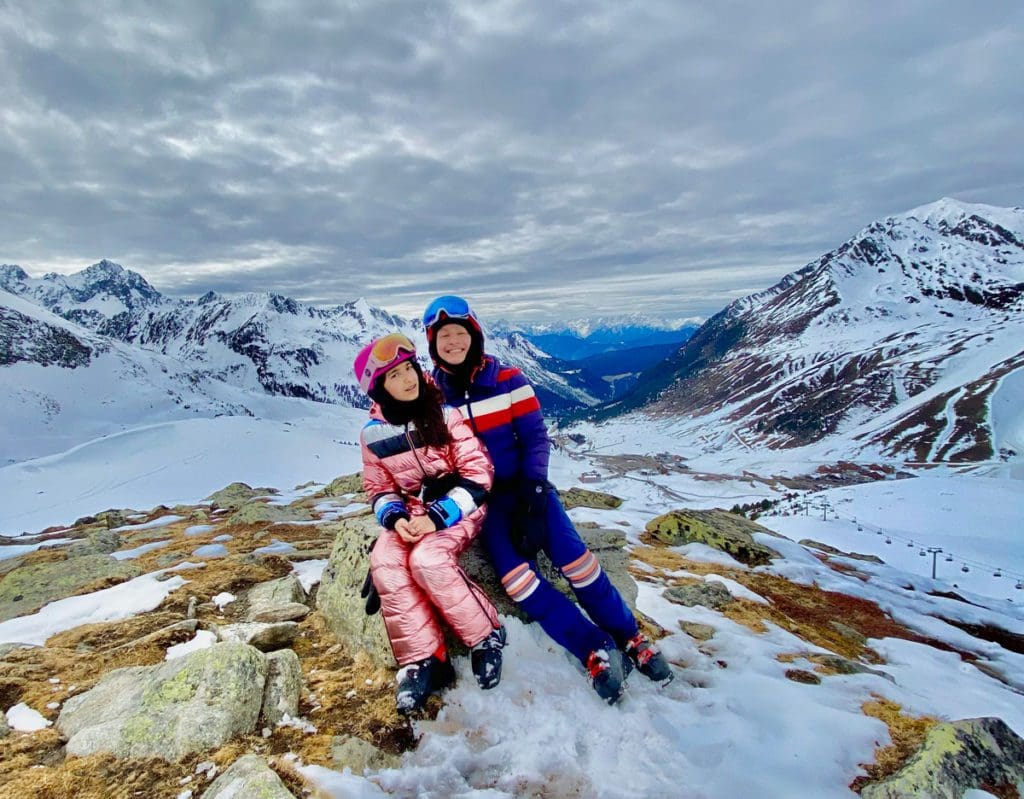 Two kids in snow gear sit on a large rock at the top of a snowy mountain, nearby ski options is just one of the many reasons why families adore Hotel Mooshaus for a family ski vacation in Austria.