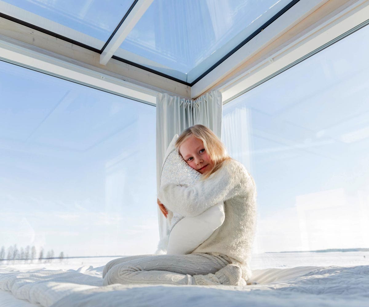 A young girl hugs a pillow, while enjoying the view from her cabin at Seaside Glass Villas, a unique accommodation option in Finland for families.