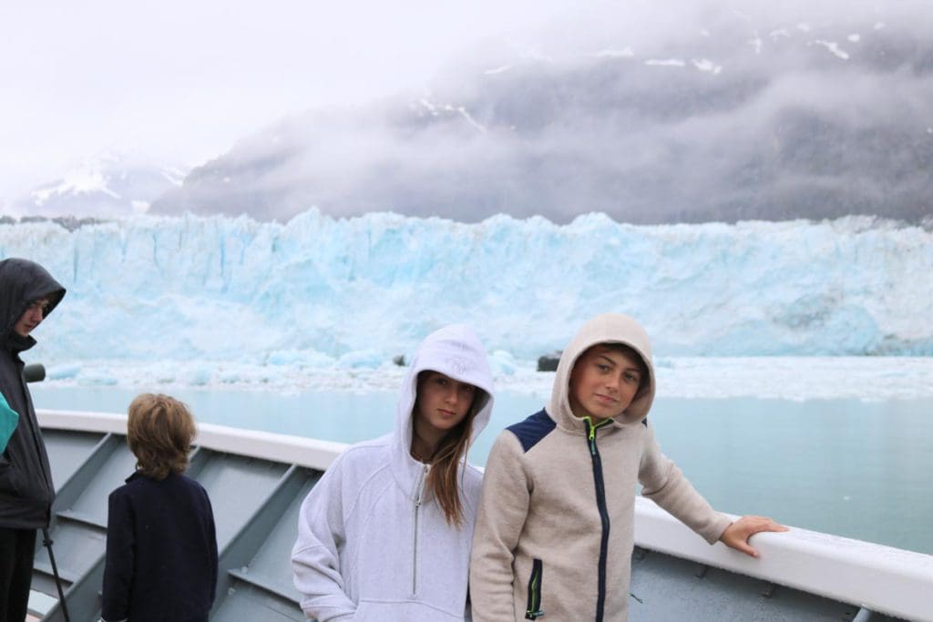Two kids stand on a cruise ship with a glacier behind them.