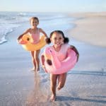 Two girls with large floaties run along a beach, enjoying a warm-weather December vacation.