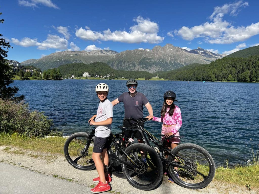 A dad and his two tweens stand together with rented bikes along the shores of Lake St. Moritz, a fun activity on any St Moritz Itinerary with kids.