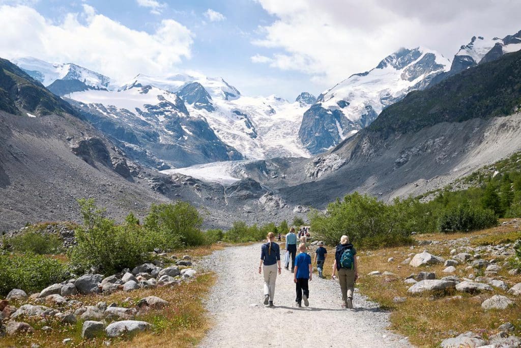 A family hikes along the Morteratsch Glacier trails in Switzerland, with mountains in the distance, a fun activity on any St Moritz Itinerary with kids.