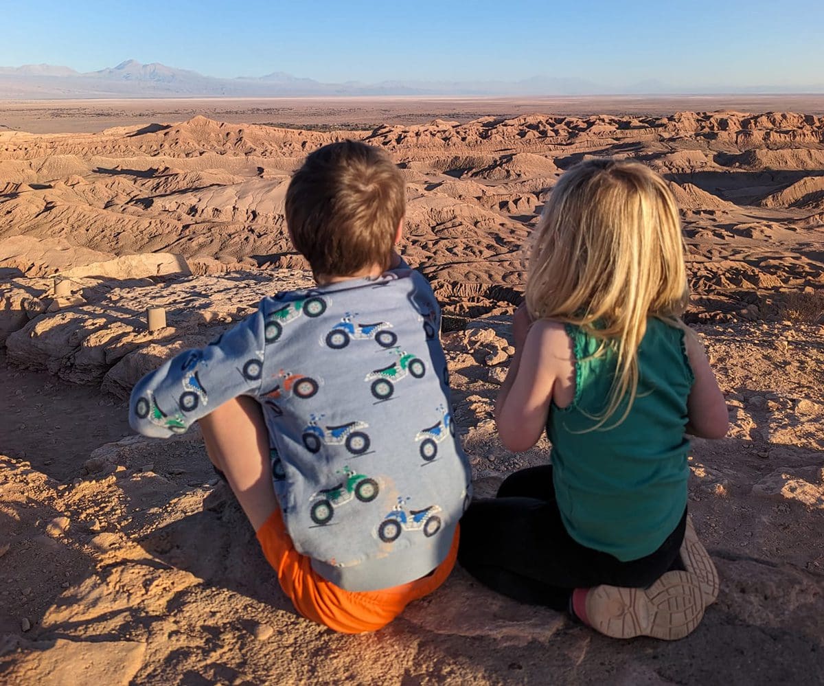 Two kids sit atop a cliff with a view of Valle de la Luna Atacama below.