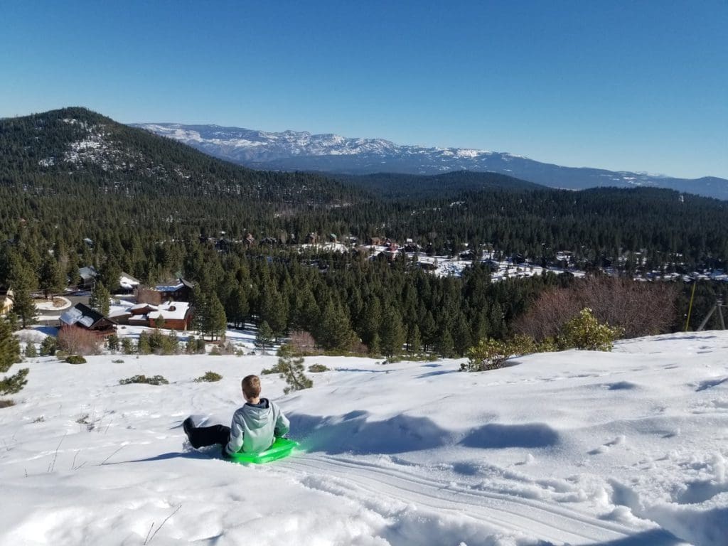 A young boy sleds down a hill on a green sled near Lake Tahoe, with mountains in the distance, one of the best things to do while on a ski vacation to Lake Tahoe with kids.