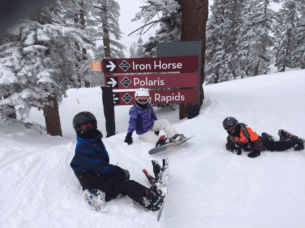 Kids sit in the snow in front of a sign showing different runs at NorthStar Resort, while on a ski vacation to Lake Tahoe with kids.