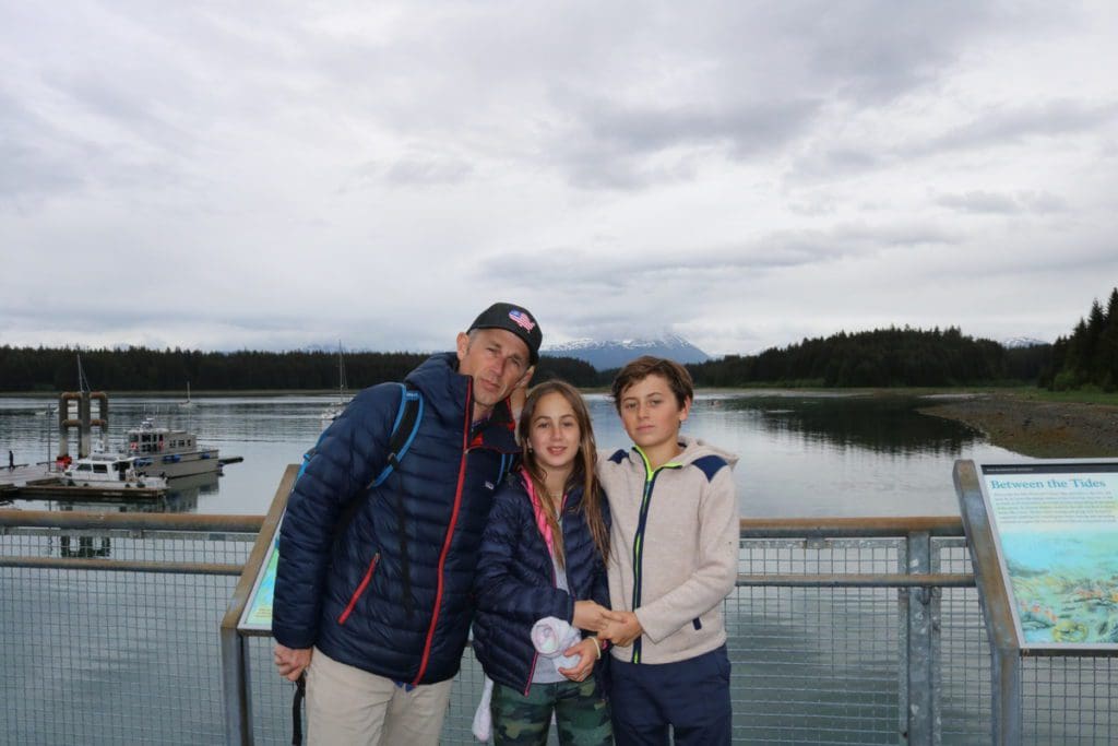 A dad and his two kids stand together, while enjoying an Alaskan cruise, with water and land in the distance.