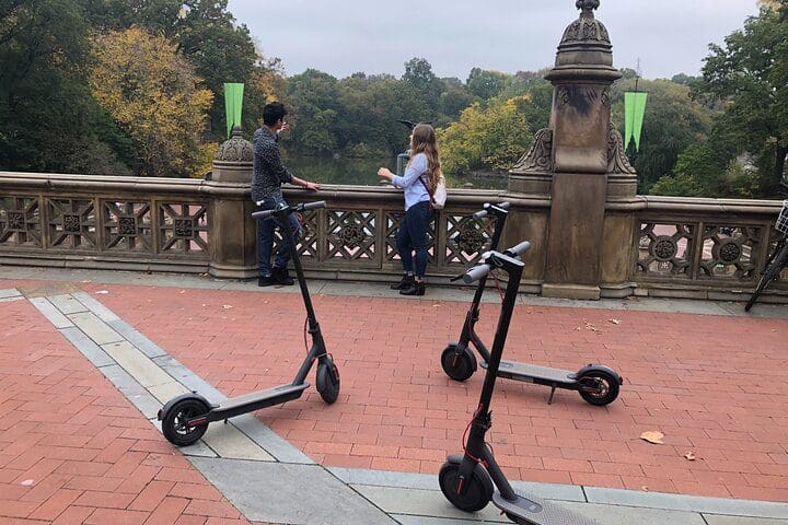 Two people stand together in Central Park with electric scooters near by awaiting the NYC Central Park Electric Scooter Tour.