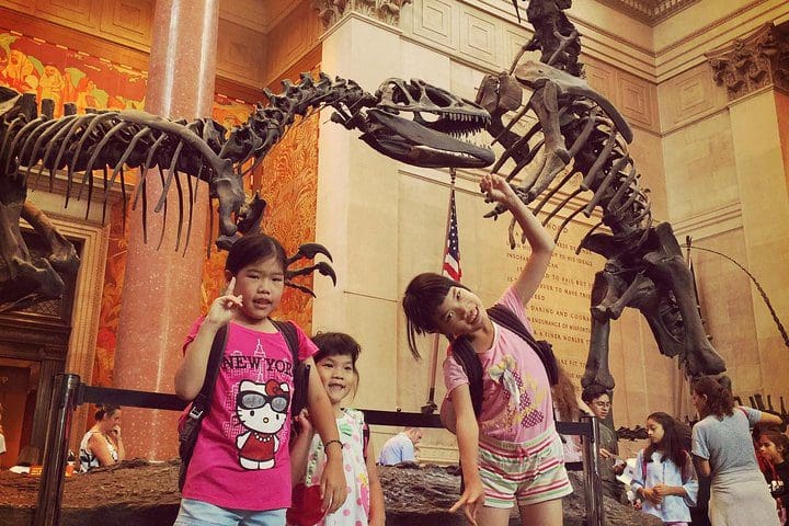 Three kids stand in front of dinosaur skeletons, while taking the NYC Kid & Families American Museum of Natural History Private Tour.