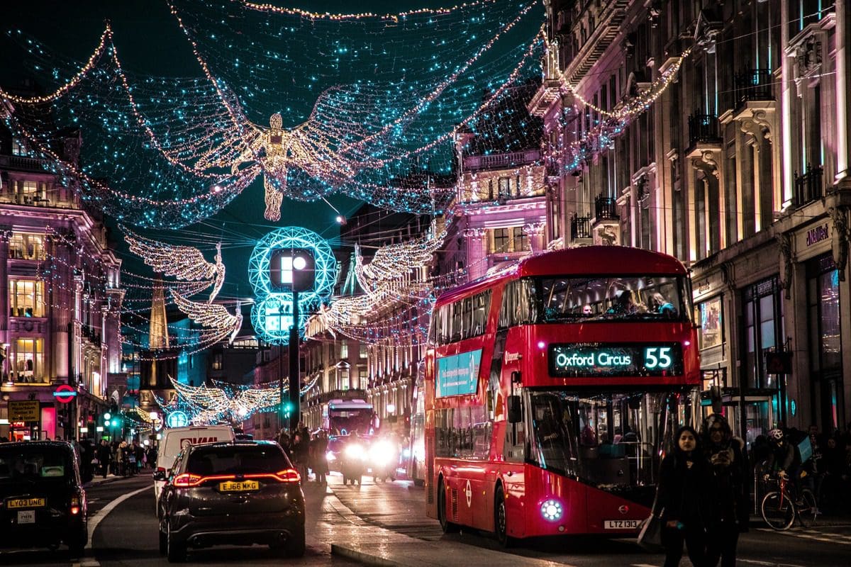 A double-decker bus moves through the streets of London, lit up with Christmas lights.