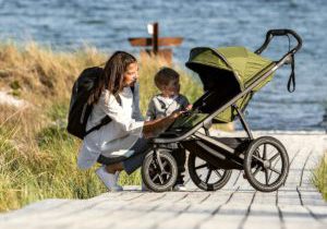 A mom kneels by her young son next to a Thule Urban Glide 2 stroller, with an ocean and tall beach grass in the distance.
