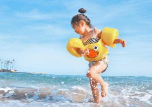 A young Asian child, wearing a duck-themed life jacket, spins in the ocean waves.