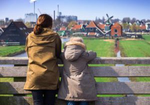 A mom and her child lean over a bridge looking at a beautiful Amsterdam scene, including greenery, a windmill, and buildings.